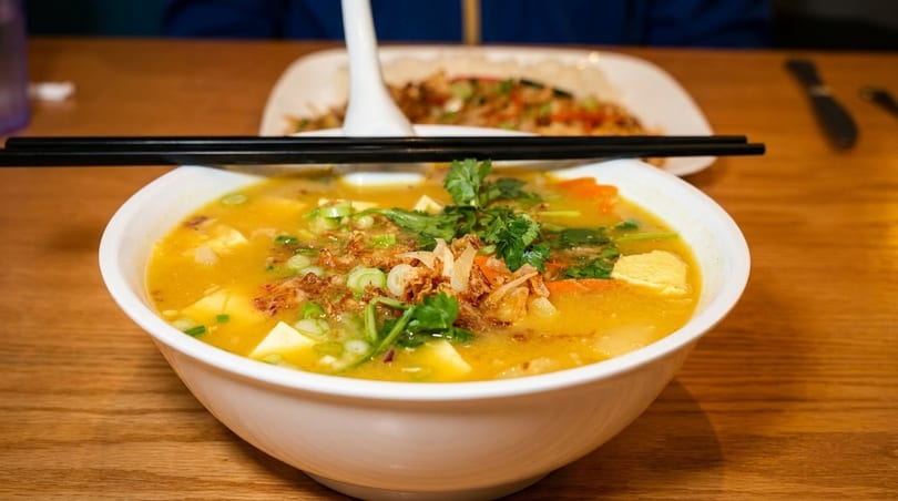 A bowl of yellow curry soup with tofu, carrots, and herbs, with chopsticks resting on top.