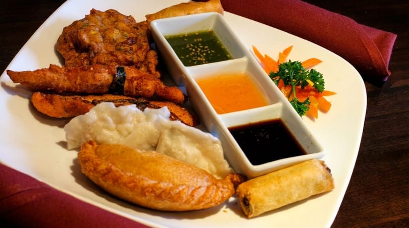 A white plate holds a variety of fried appetizers, dumplings, and three dipping sauces.